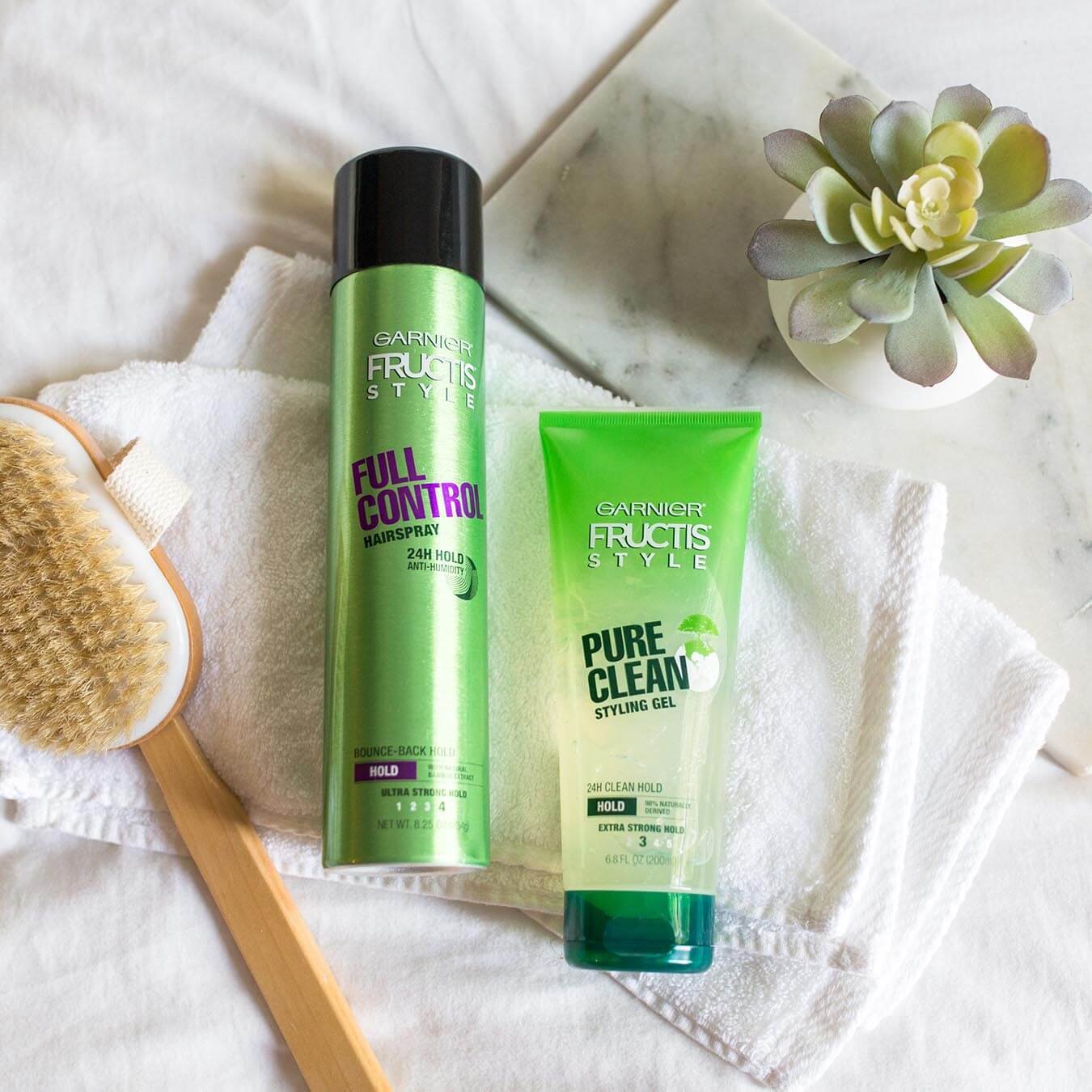 Garnier Fructis Style Full Control Hairspray and Fructis Style Pure Clean Styling Gel on two white wash cloths next to a wooden long-handled scrub brush and a potted succulent.