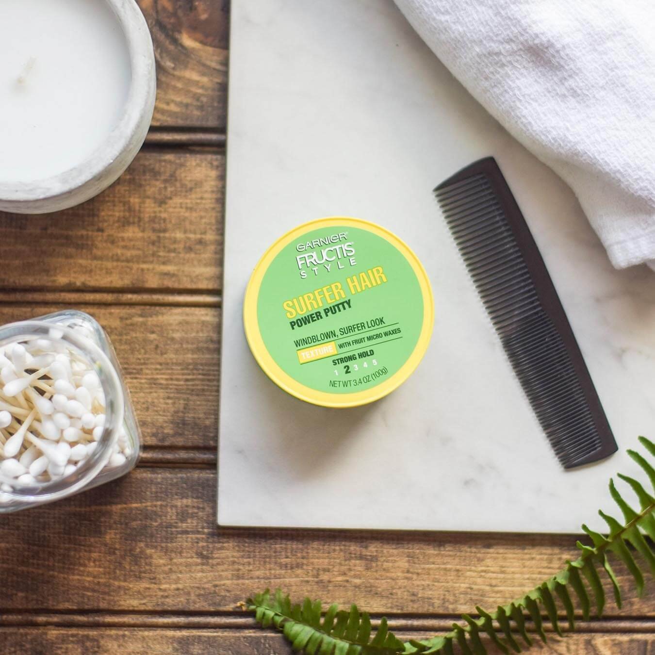 Garnier Fructis Style Surfer Hair Power Putty on a white marble slab with a black comb and white hand towel on a wooden table with a white candle, jar of q-tips, and fern frond.
