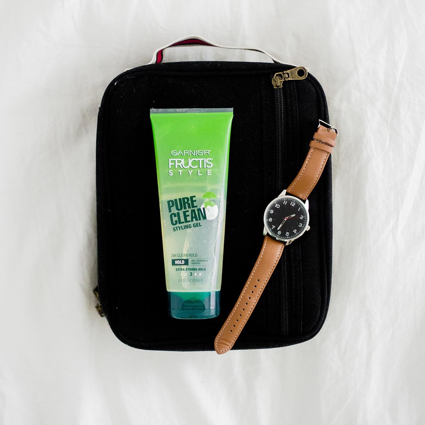Garnier Fructis Style Pure Clean Styling Gel next to a watch with a black face and brown band on a black travel bag on white cloth.