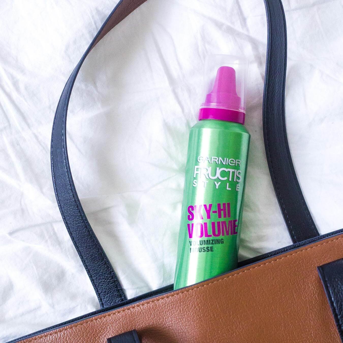 Garnier Fructis Style Sky-Hi Volume Volumizing Mousse peeking out of a brown purse with black straps on white cloth.