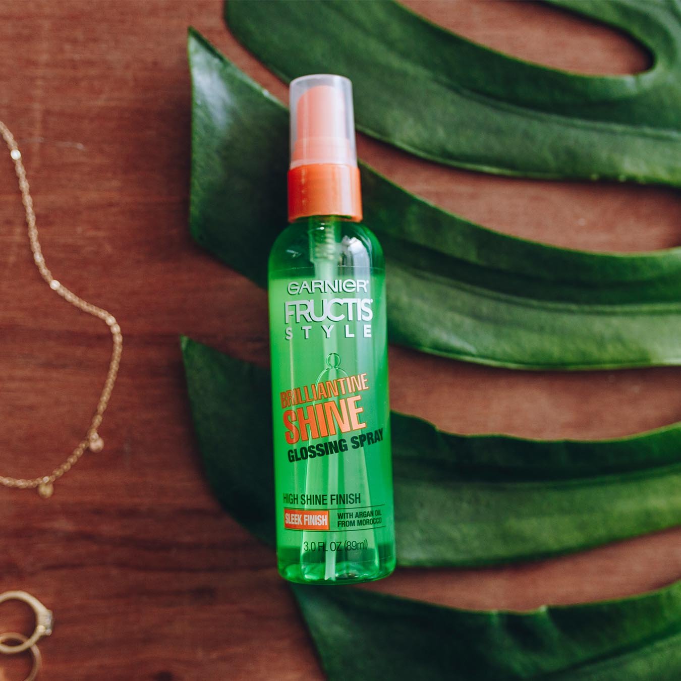 Garnier Fructis Style Brilliantine Shine Glossing Spray on a palm frond on a warm wooden table next to a gold bracelet and two gold rings.