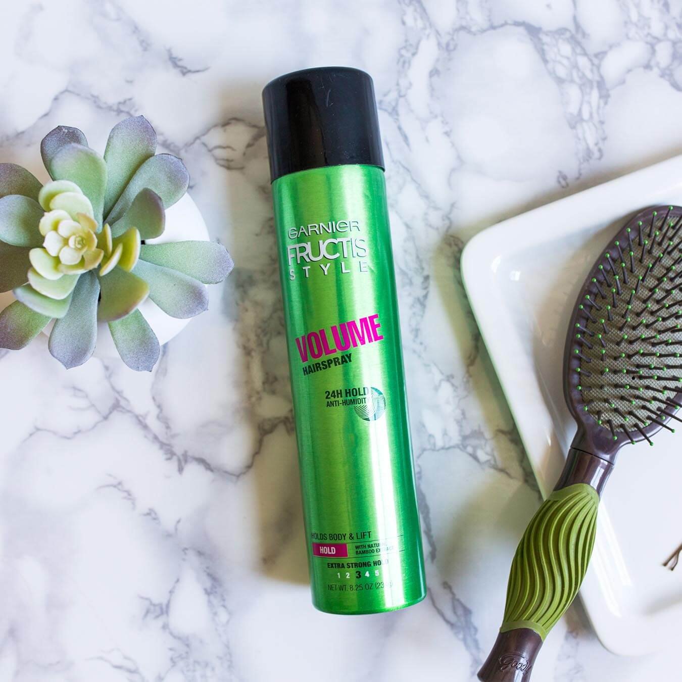 Garnier Fructis Style Volume Hairspray on white marble next to a potted succulent and a white tray holding a bobby pin and a green and brown brush.