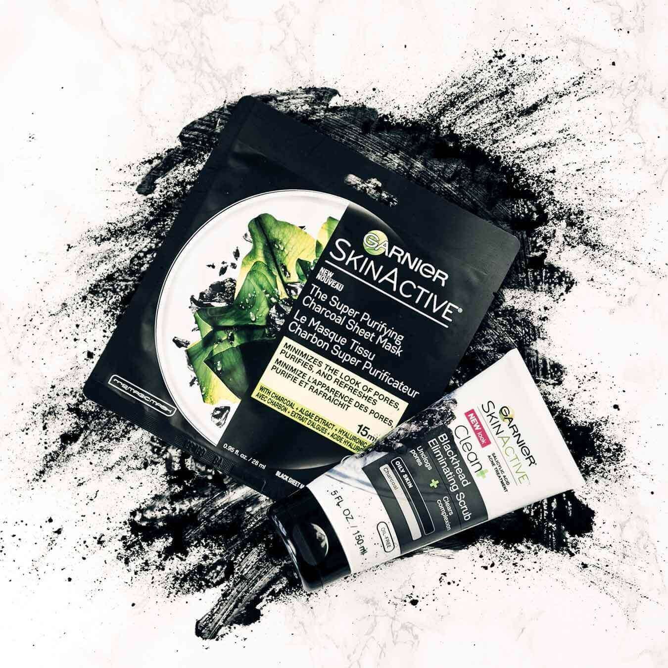 Garnier SkinActive Super Purifying Charcoal Sheet Mask and SkinActive Clean+ Blackhead Eliminating Scrub on an explosion of charcoal powder on pinkish white marble.