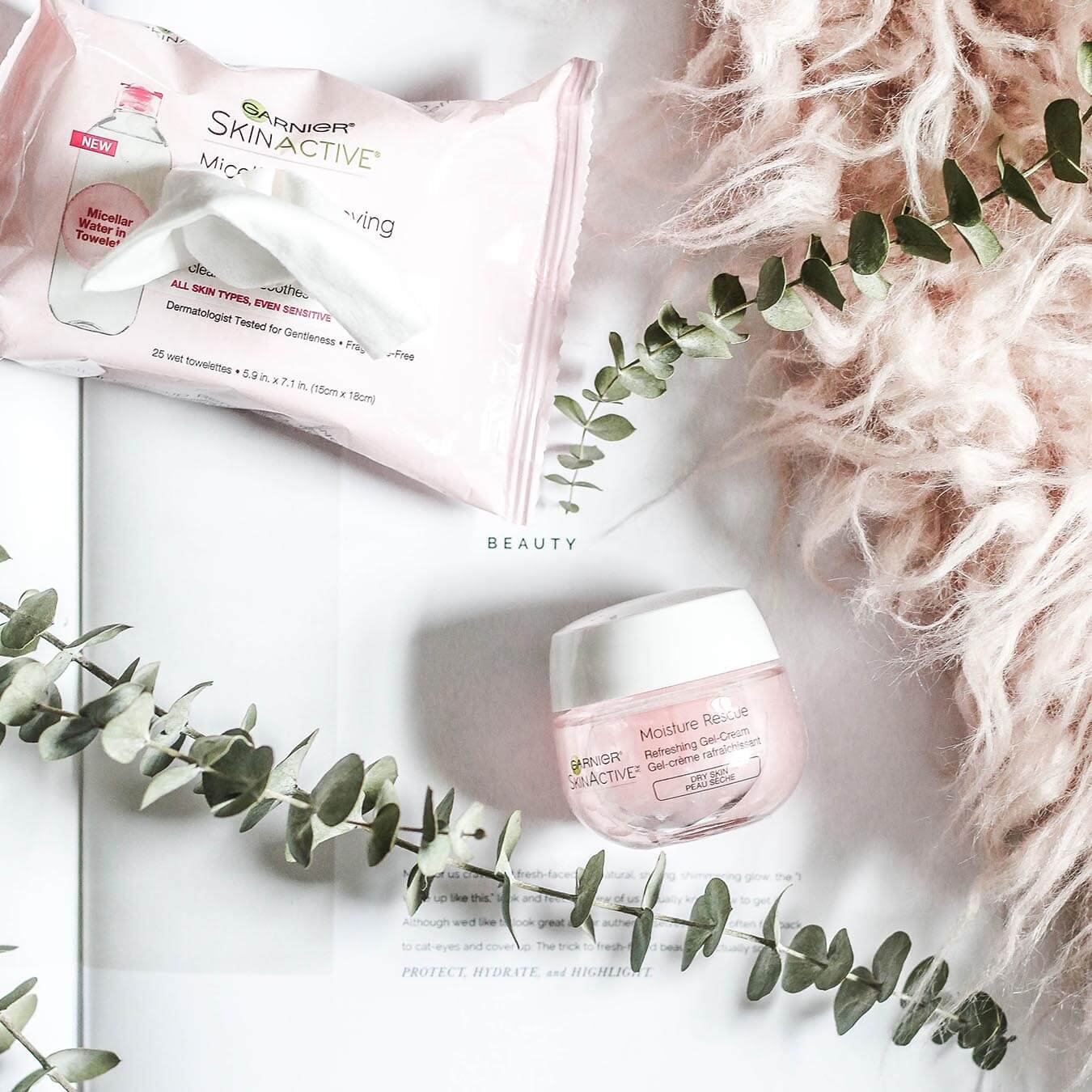 Garnier SkinActive Moisture Rescue Refreshing Gel-Cream laying on its side and an opened SkinActive Micellar Makeup Removing Towelettes All-in-1 laying on an open magazine partially covered by eucalyptus leaves and a pink shag rug revealing the word "beauty" on the page.