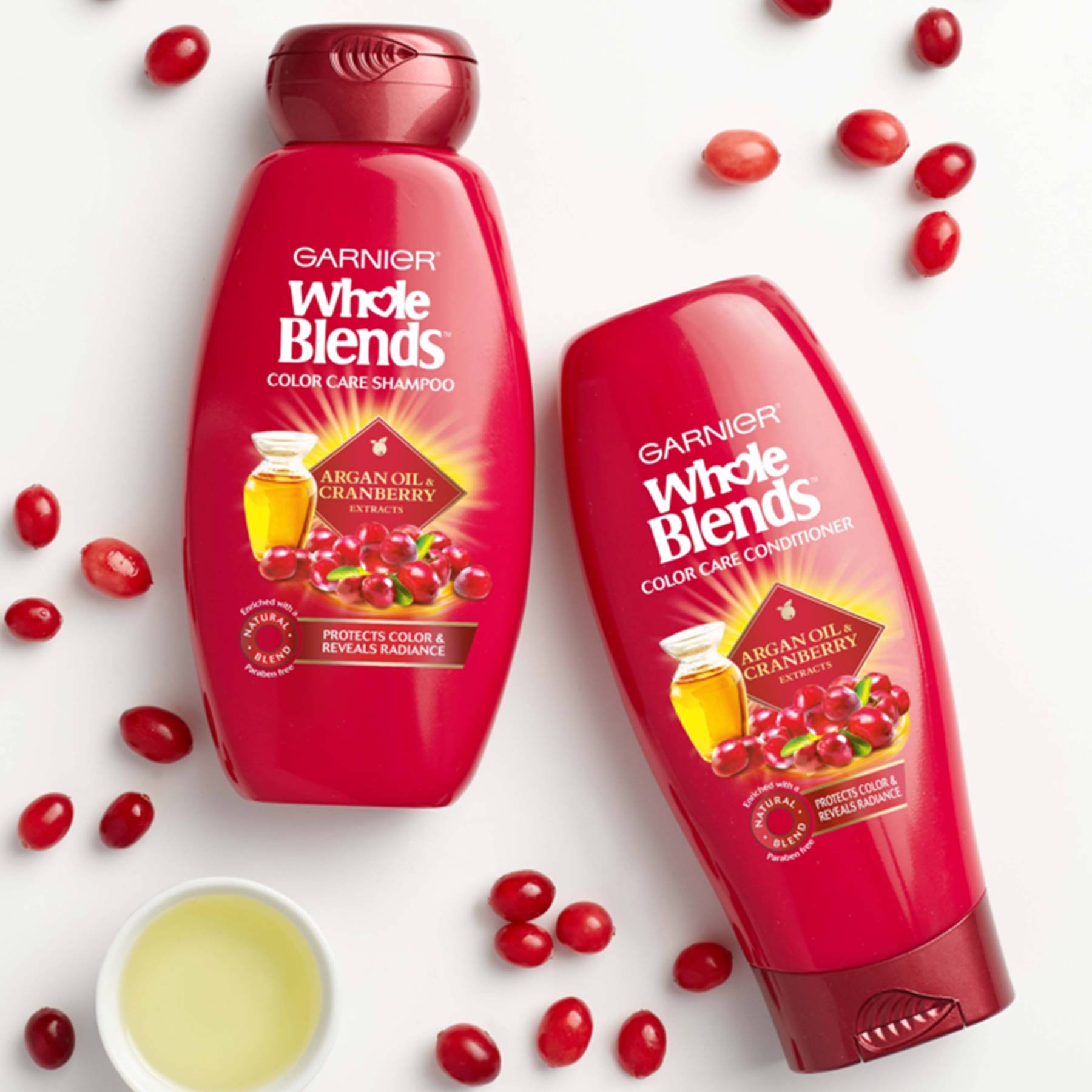 Whole Blends Color Care Shampoo with Argan Oil and Cranberry and Color Care Conditioner with Argan Oil and Cranberry on a white background strewn with whole cranberries and a ramekin of oil.