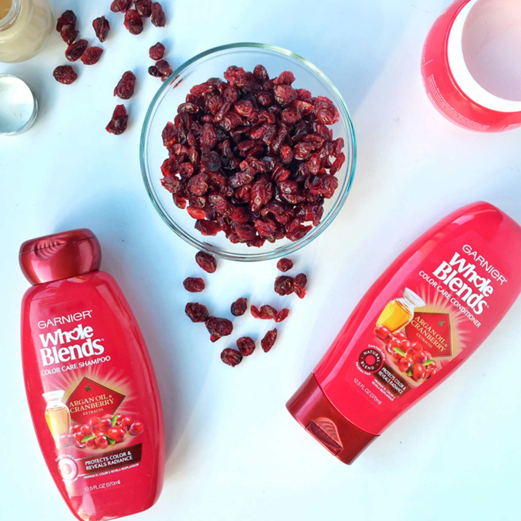 Whole Blends Color Care Shampoo with Argan Oil and Cranberry and Color Care Conditioner with Argan Oil and Cranberry on a blue-white background with an open jar of Argan Oil and Cranberry Color Care Mask, open jar of foundation, and a bowl of dried cranberries with some spilled out.