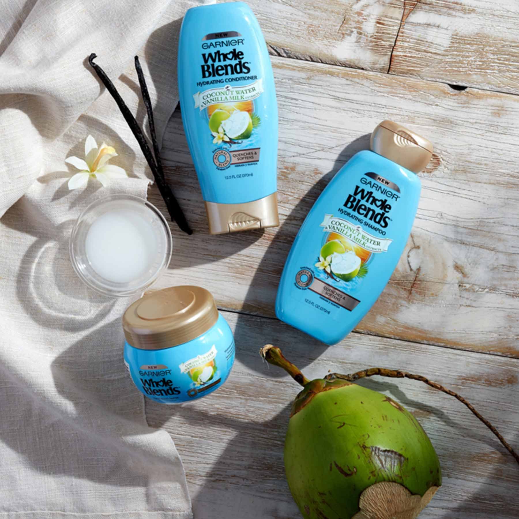 Whole Blends Coconut Water and Vanilla Milk Shampoo, Coconut Water and Vanilla Milk Conditioner, and Coconut Water and Vanilla Milk Hydrating Mask on distressed wooden planks next to glass ramekin of coconut oil, two vanilla beans, a vanilla flower, a green coconut on its branch, and some natural colored linen.