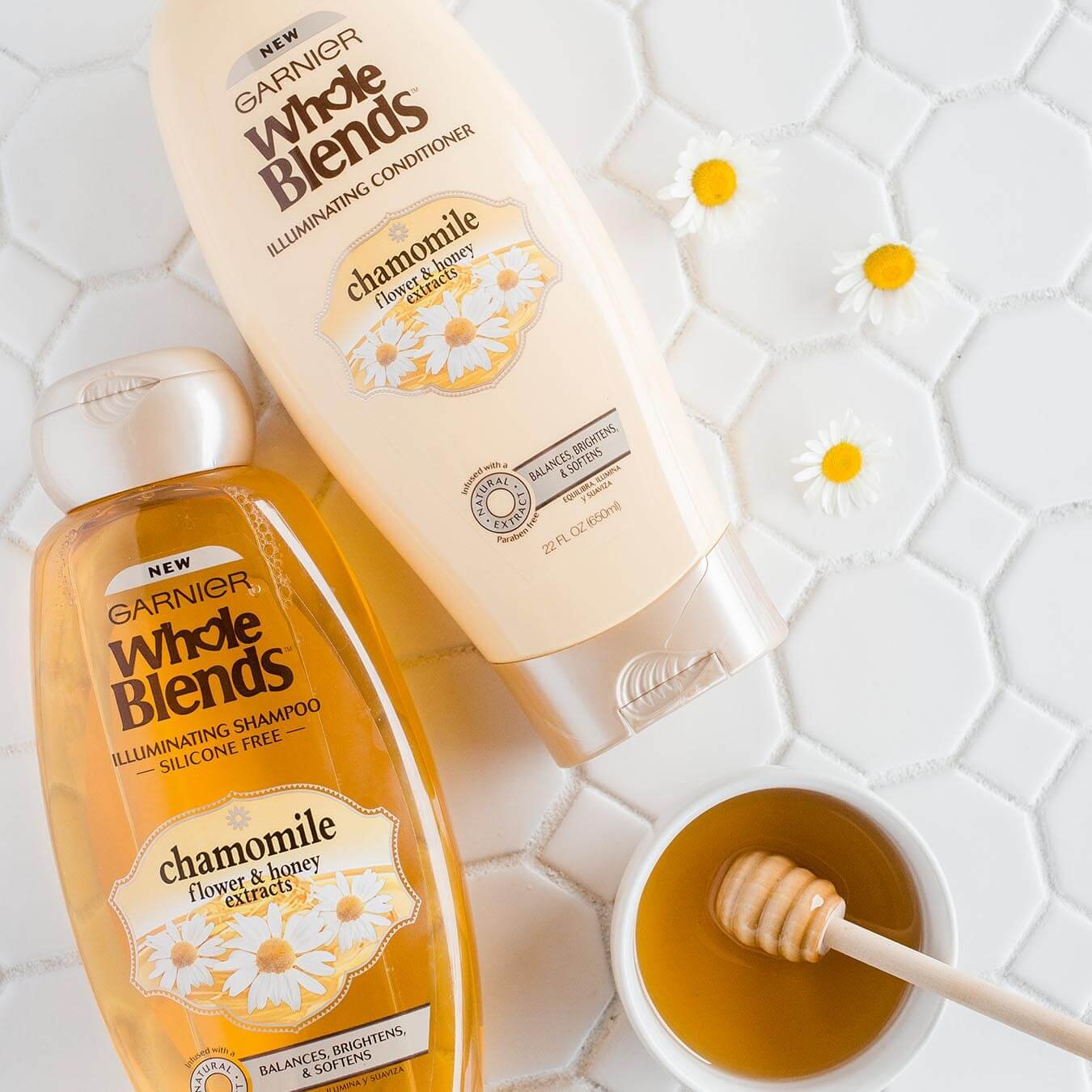 Whole Blends Chamomile Illuminating Shampoo with Flower and Honey Extracts and Chamomile Illuminating Conditioner with Flower and Honey Extracts on a background of octagonal and diamond tiles strew with daisy blossoms and a ramekin of honey with a honey dipper.