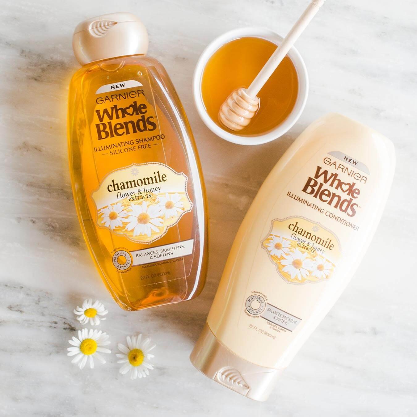 Whole Blends Chamomile Illuminating Shampoo with Flower and Honey Extracts and Chamomile Illuminating Conditioner with Flower and Honey Extracts on white marble with daisy blossoms and a ramekin of honey with a honey dipper.