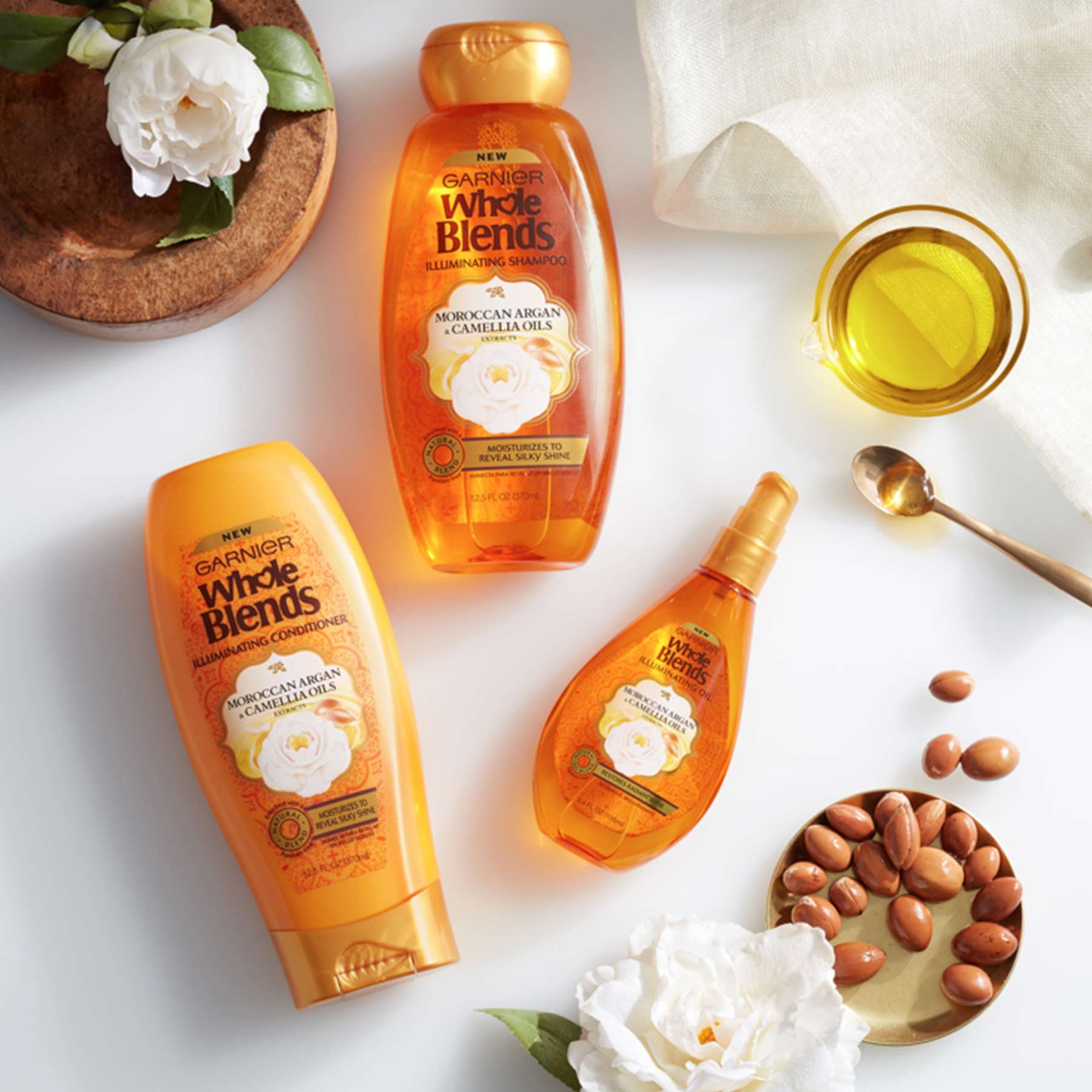 Whole Blends Illuminating Moroccan, Argan, & Camellia Oils Shampoo, Illuminating Moroccan, Argan, & Camellia Oils Conditioner, and Illuminating Moroccan, Argan, & Camellia Oils Illuminating Oil on a white background with white linen, a glass of oil, a bronze teaspoon with oil, a bronze tin of argan nuts, and two camellia flowers, one of which is in a thick wooden bowl.