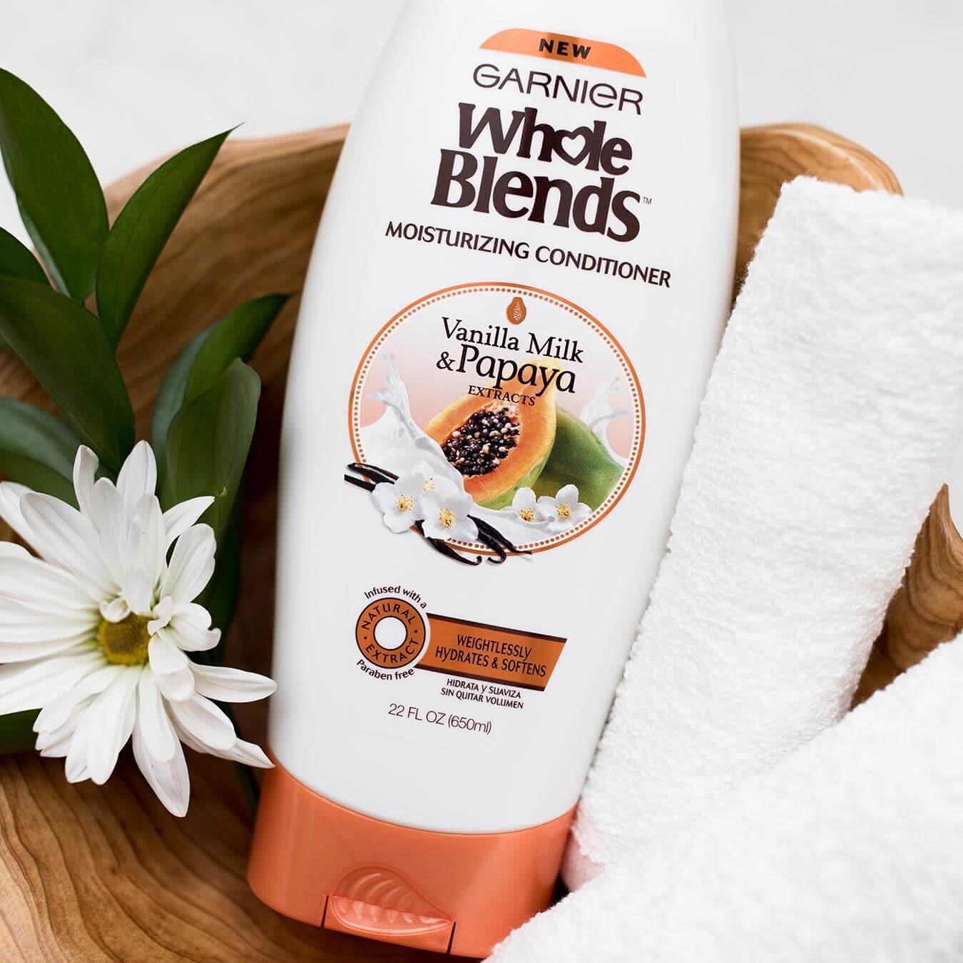 Whole Blends Vanilla Milk and Papaya Moisturizing Conditioner in an irregular wooden bowl next to some leaves, a daisy blossom, and two rolled white hand towels.