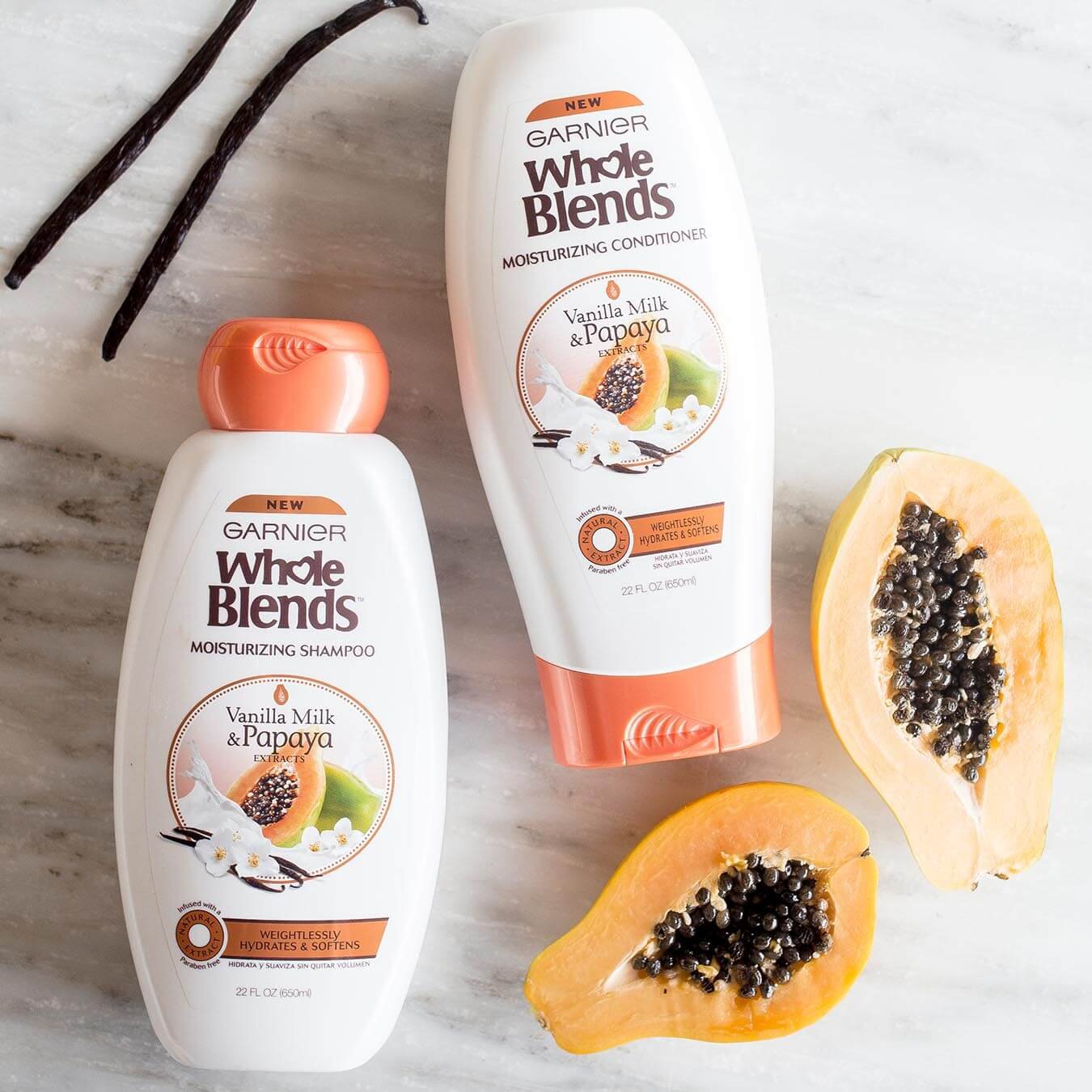 Whole Blends Vanilla Milk and Papaya Moisturizing Shampoo and Vanilla Milk and Papaya Moisturizing Conditioner on white marble next to a halved papaya and two vanilla beans.