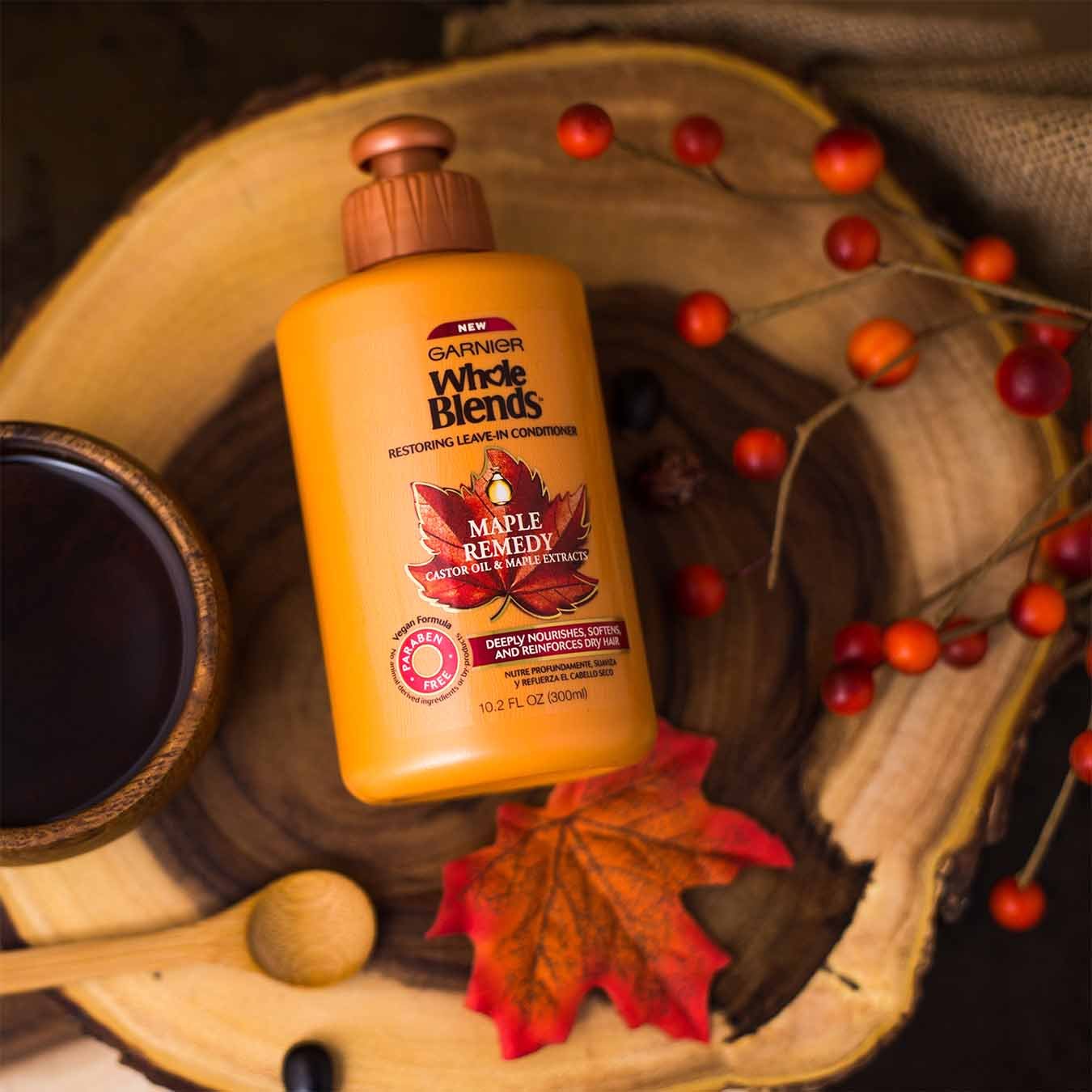 Whole Blends Maple Remedy Leave-In Conditioner with Caster Oil and Maple Extracts falling into a wooden bowl that contains a leafless bough of cranberries, a small wooden bowl of maple syrup, a wooden spoon, and a red-orange maple leaf on a woven cloth and wooden table.