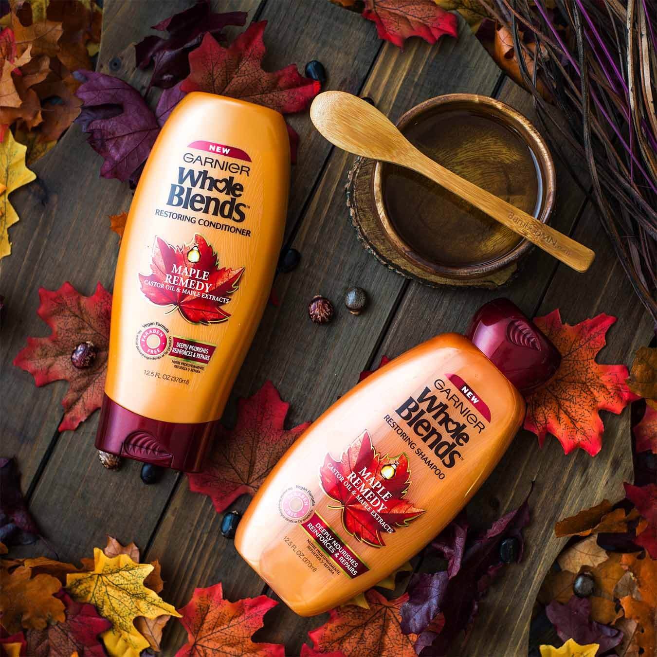 Whole Blends Maple Remedy Shampoo with Caster Oil and Maple Extracts and Maple Remedy Conditioner with Caster Oil and Maple Extracts next to a wooden bowl of maple syrup with a wooden spoon on top beside a woven tangle of branches and purple wires on a wooden table strewn with red, orange, and purple maple leaves and beads.