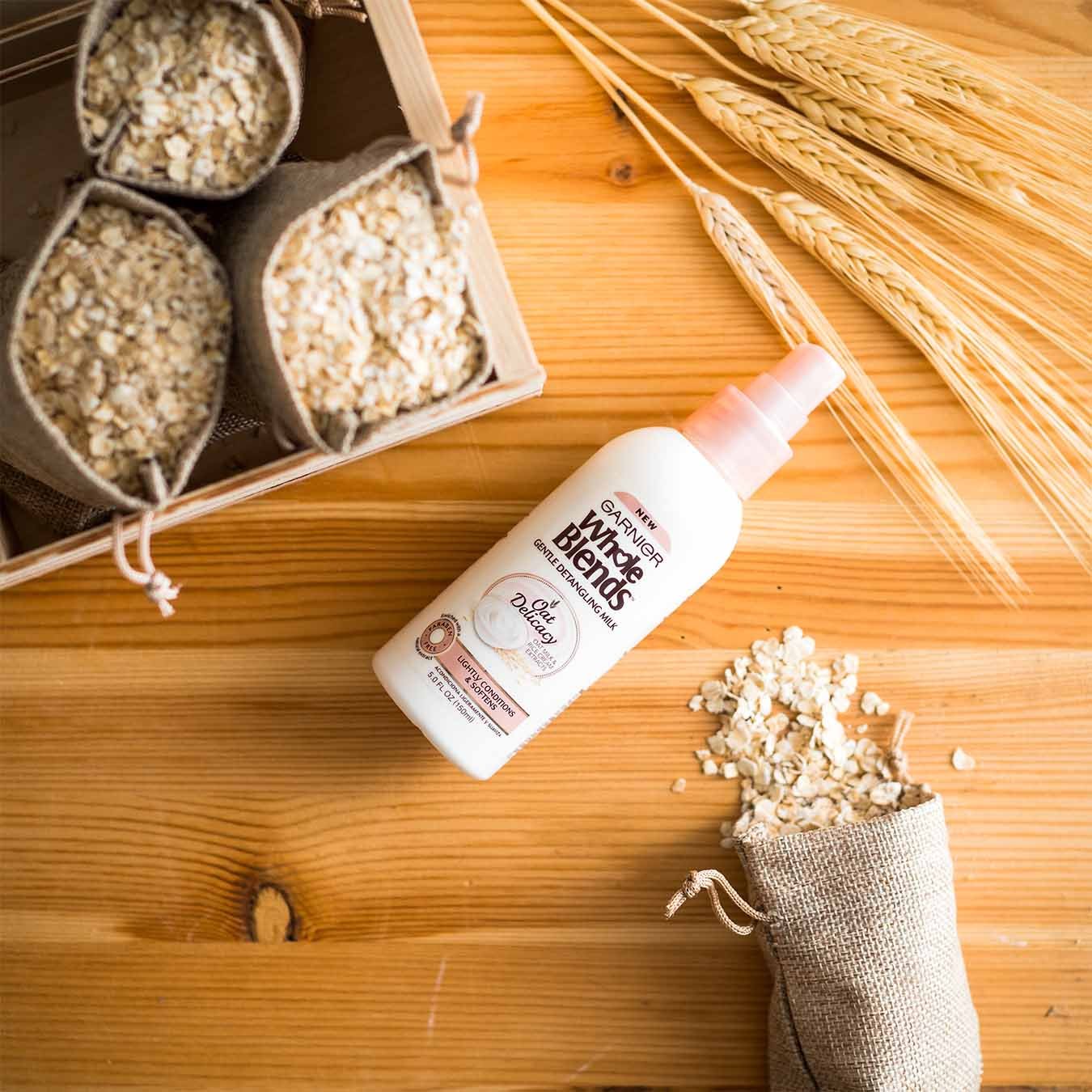 Whole Blends Oat Delicacy Gentle Detangling Milk on a wooden table with wheat stalks and a crate of small burlap sacks of oats, one of which has spilled.
