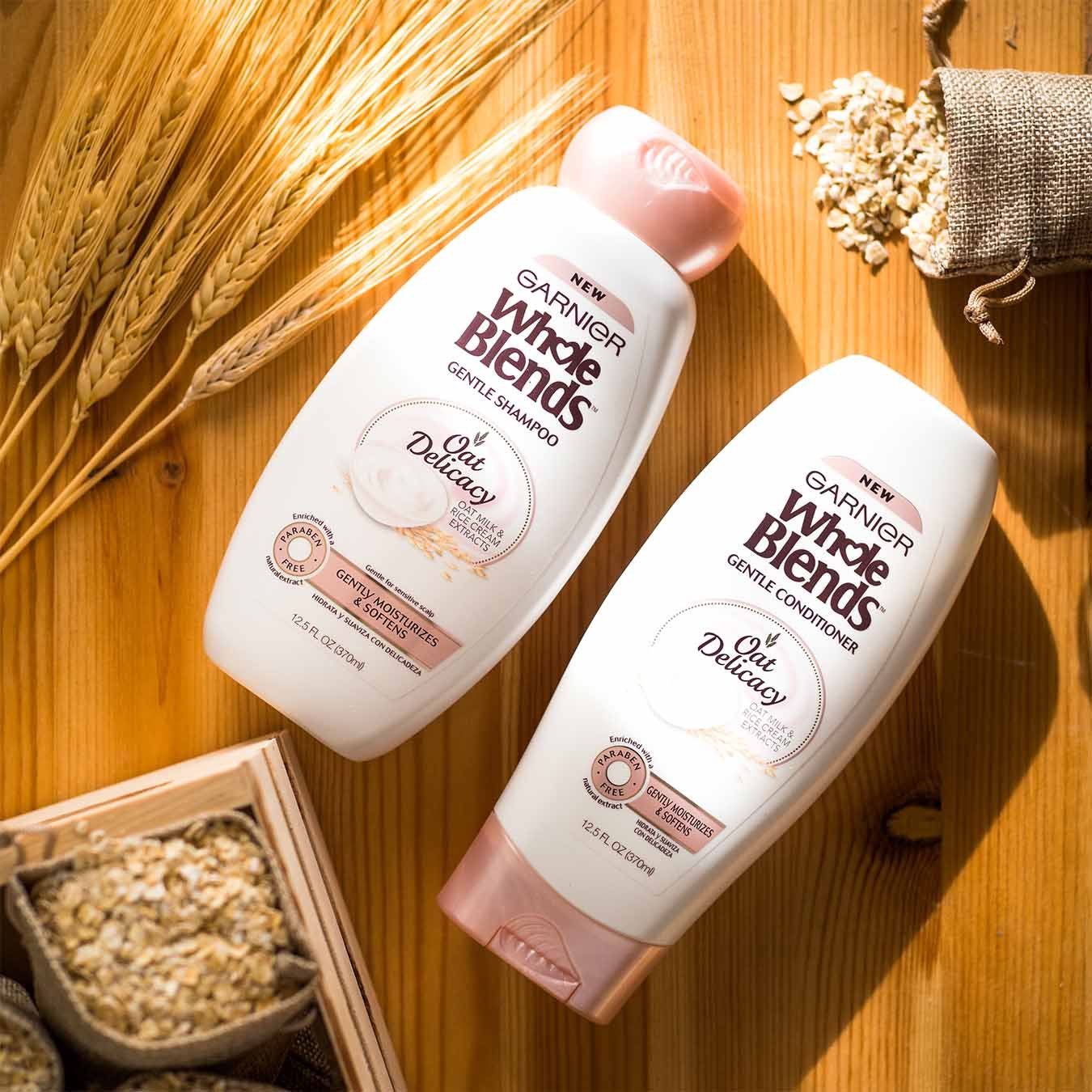 Whole Blends Oat Delicacy Shampoo and Oat Delicacy Conditioner on a wooden table with wheat stalks and a crate of small burlap sacks of oats, one of which has spilled.