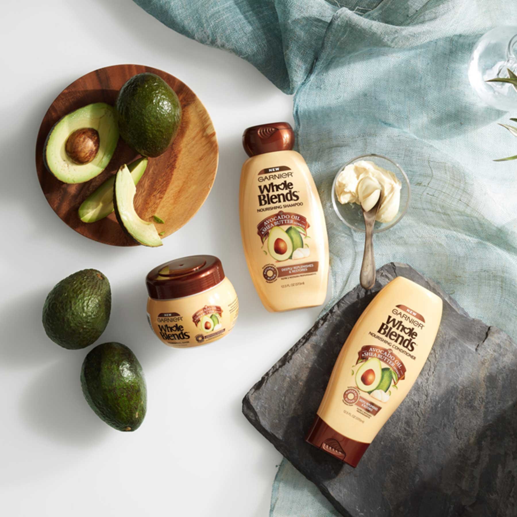 Whole Blends Avocado Oil and Shea Butter Shampoo, Avocado Oil and Shea Butter Conditioner, and Avocado Oil and Shea Butter Nourishing mask next to a slab of slate, glass vase with a leafy plant, a round wooden slab of sliced avocado, whole avocados, and faded blue linen on a white background.