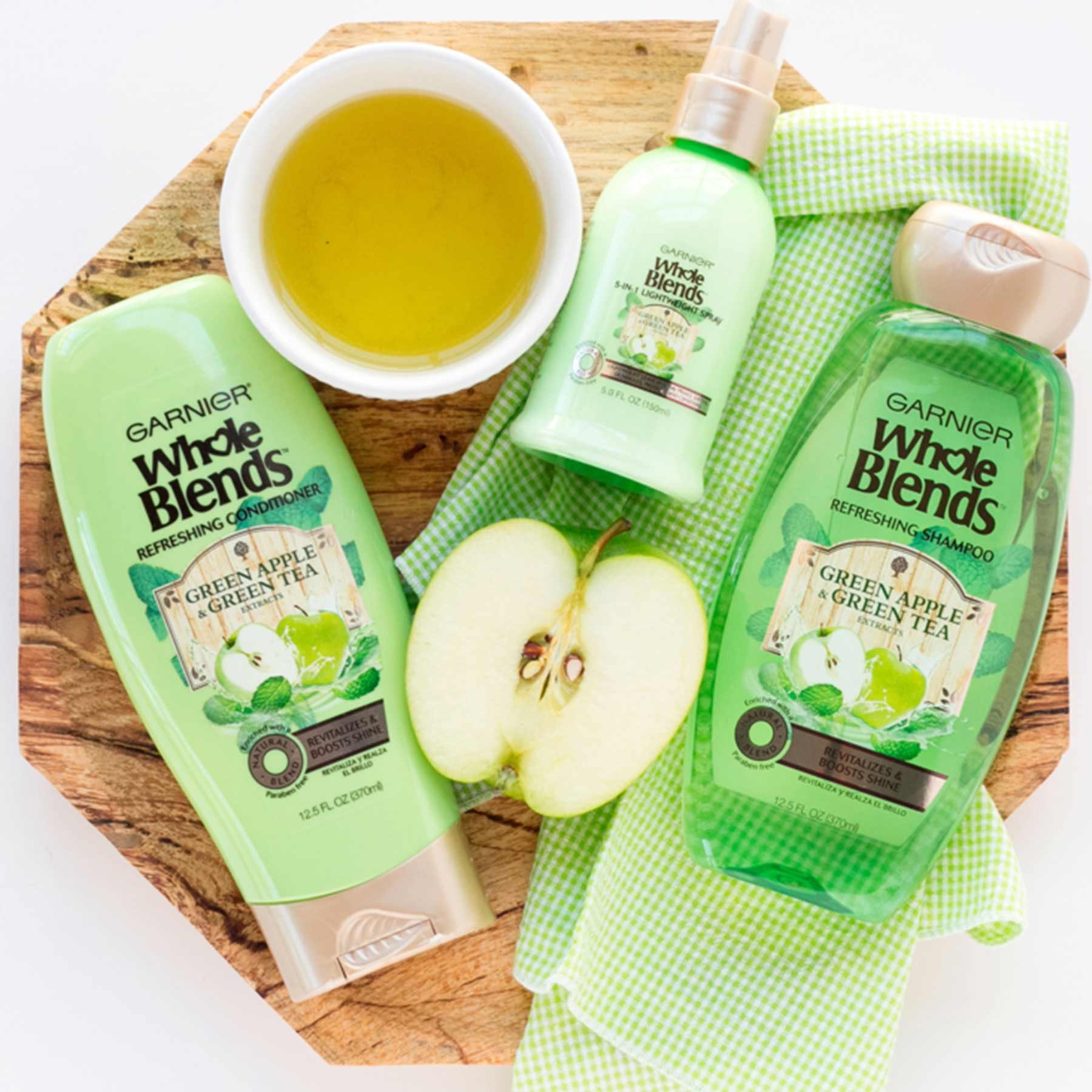 Whole Blends Green Apple and Green Tea Shampoo, Green Apple and Green Tea Conditioner, and Green Apple and Green Tea 5-in-1 Lightweight Spray on an octagonal wooden board with a white cup of green tea, half of a granny smith apple, and a green and white checkered cloth napkin on a white background.