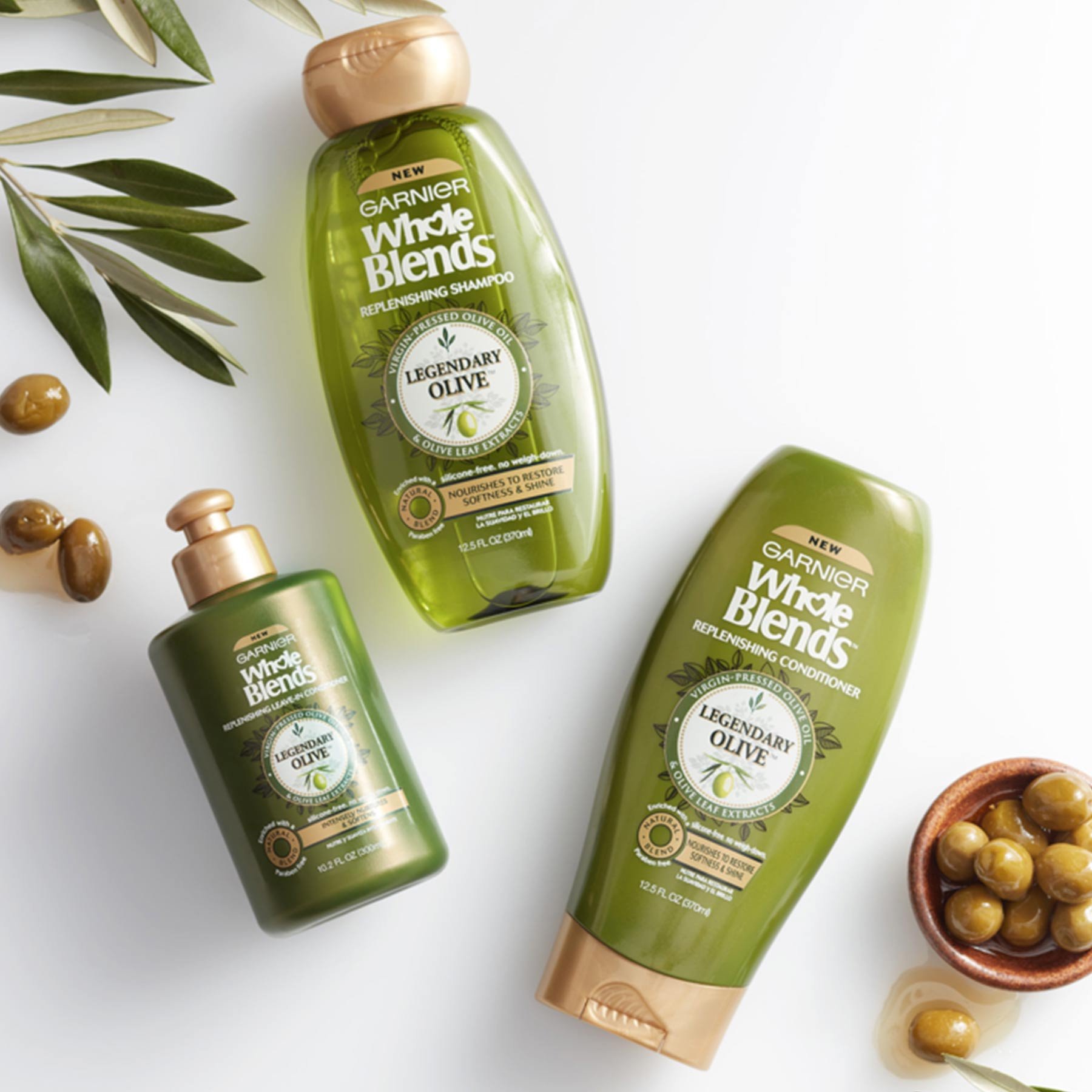 Whole Blends Legendary Olive Shampoo, Legendary Olive Conditioner, and Legendary Olive Leave-In Conditioner on a white background next to an earthen bowl of green olives in olive juice with some spilled out and olive leaves.