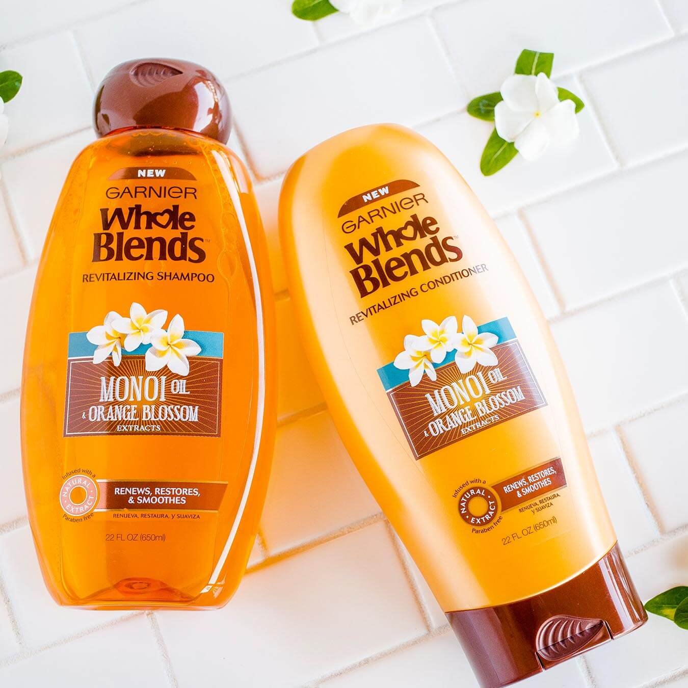 Whole Blends Monoi Oil Shampoo with Orange Blossom Extract and Monoi Oil Conditioner with Orange Blossom Extract on white tiles strewn with monoi blossoms.