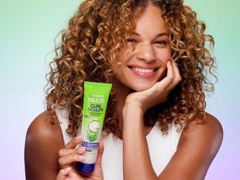 Woman with 3B curls smiling at the camera while holding up a bottle of Garnier Sculpt Conditioning Cream Gel.