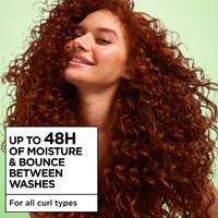 A smiling woman with long, voluminous red curly hair and freckles poses against a green background beside text reading "Up to 48H of Moisture & Bounce Between Washes, for all curl types."