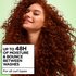 A smiling woman with long, voluminous red curly hair and freckles poses against a green background beside text reading "Up to 48H of Moisture & Bounce Between Washes, for all curl types."