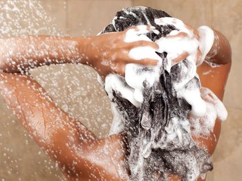 The back of a person's head and shoulders in a shower, with hands vigorously shampooing dark, lathered hair under a stream of water.