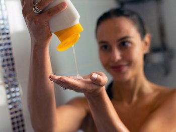 A blurred image of a person pouring white shampoo from a yellow-capped bottle into their hand in a shower.