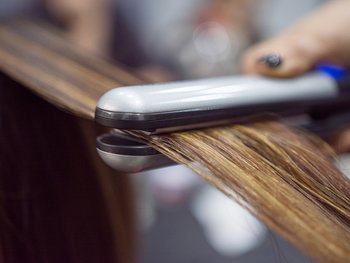 A close-up of a flat iron straightening a section of brown hair with blonde highlights.
