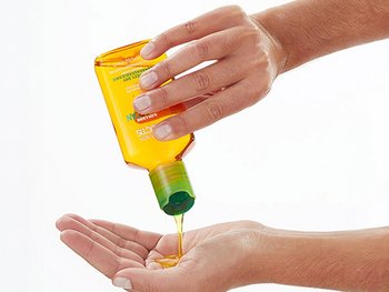 Hand tilting a yellow bottle with a green cap to pour golden hair oil into an open palm.