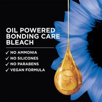 Dark navy promotional graphic for Garnier Olia featuring a large, glistening golden oil droplet at center-right with soft blue flowers in the background. Bold white headline text reads: "OIL POWERED BONDING CARE BLEACH." Below, a bulleted checklist in white reads: "✓ No Ammonia ✓ No Silicones ✓ No Parabens ✓ Vegan Formula."