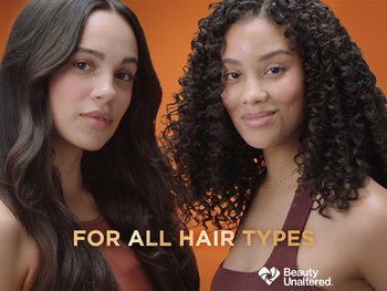 Two women smiling naturally at the camera – one has long, smooth, wavy brown hair, and the other has 3C brown hair. Honey Milk is for all hair types.