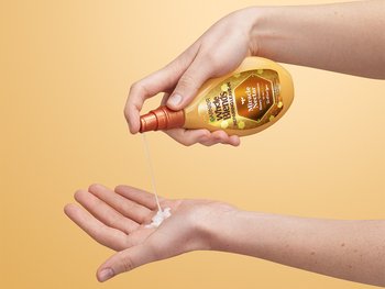 Hands dispensing a white, creamy product from a bottle of Garnier Whole Blends Miracle Nectar Honey Treasures 10-in-1 Repairing Leave-In into a palm.