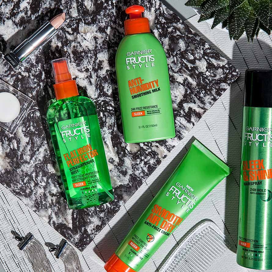 Anti-Humidity Smoothing Milk, Sleek & Shine Flat Iron Perfector Straightening Mist, Smooth Air Dry Anti-Frizz Cream, and Sleek & Shine Anti-Humidity Aerosol Hairspray on a black and white mat, with a plant, a pink lipstick, silver earrings, and a transparent comb.