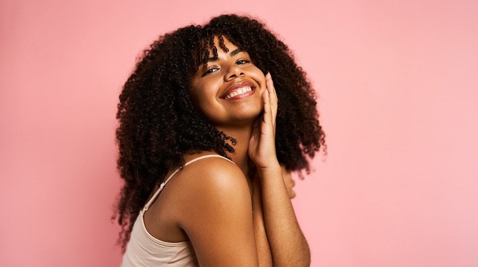 A person with dark skin and curly hair smiles and touches their face.