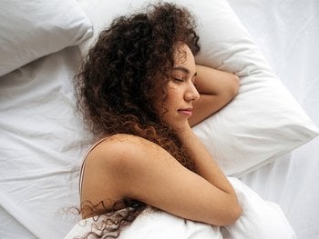 A person with curly hair is peacefully sleeping on a white bed with their head on a pillow.