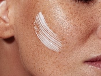 Close-up of a person's cheek with visible freckles and a streak of white cream applied to the skin.