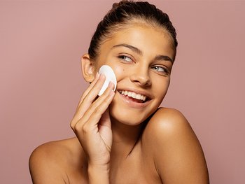 Teenage girl smiling as she uses a toner pad to cleanser her face.