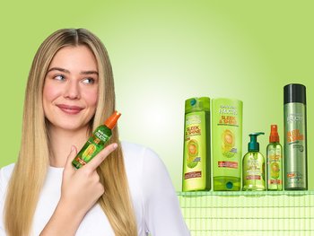 Blonde female with sleek straight hair down to her sides. She's looking up to the side as she smiles and holds a bottle of Sleek and Shine's Brilliantine Shine Spray.