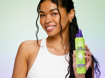 Woman with long 2B wavy curls facing the camera holding up a bottle of Garnier Construct Mousse.